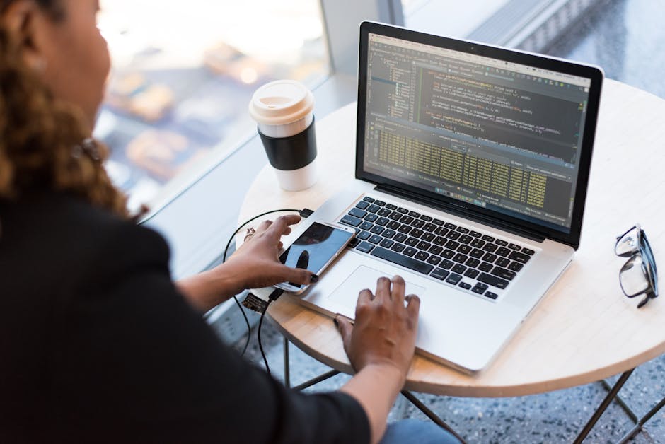 Black woman programming on a laptop with coffee, smartphone, and glasses on a desk in an office